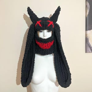 May include: Black knitted hat with long bunny ears and a red stitched smile. The hat has a black knitted face mask with red X's for eyes.