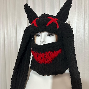 May include: Black knitted hat with long, floppy ears and a red stitched design resembling a face with horns.