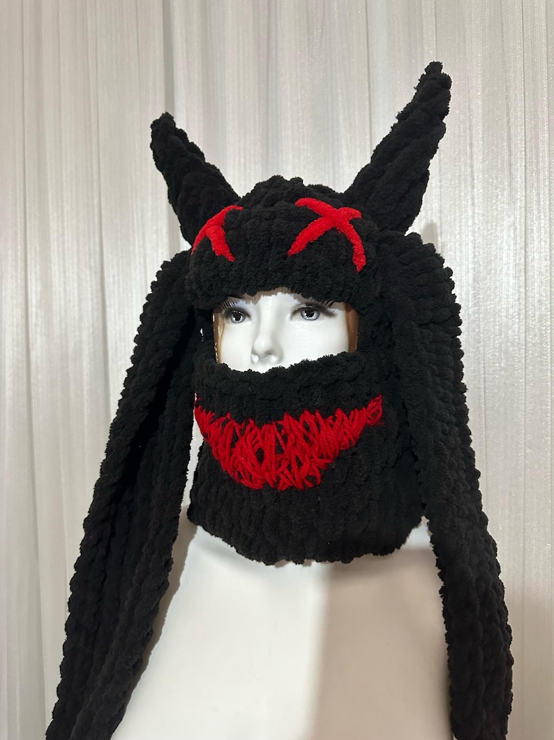 May include: Black knitted hat with long, floppy ears and a red stitched design resembling a face with horns.