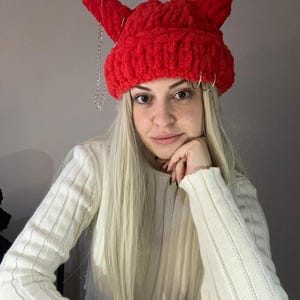 Pode incluir: Um gorro vermelho de croch&ecirc; com chifres de diabo e uma corrente prateada. O gorro &eacute; usado por uma pessoa com cabelo longo e loiro.