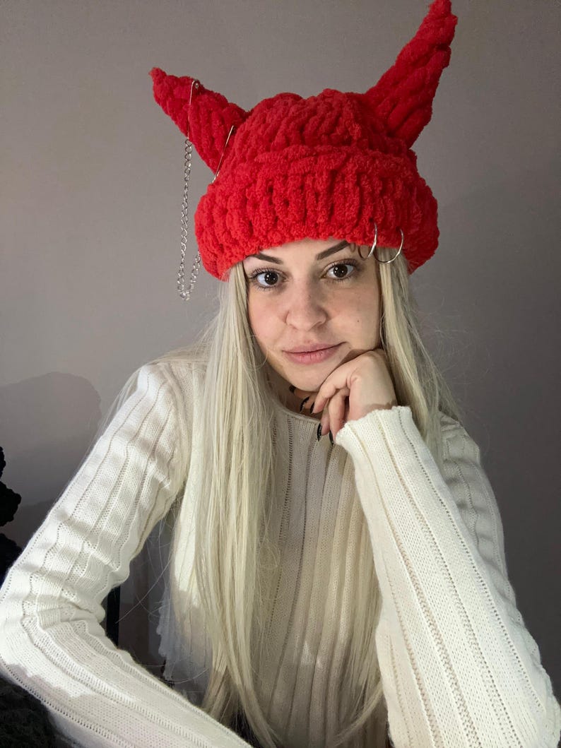 Pode incluir: Um gorro vermelho de croch&ecirc; com chifres de diabo e uma corrente prateada. O gorro &eacute; usado por uma pessoa com cabelo longo e loiro.