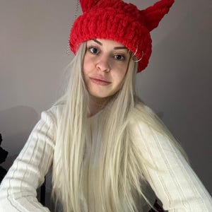 Pode incluir: Um gorro vermelho de croch&ecirc; com chifres de diabo e uma corrente prateada. O gorro &eacute; usado por uma pessoa com cabelo longo e loiro.