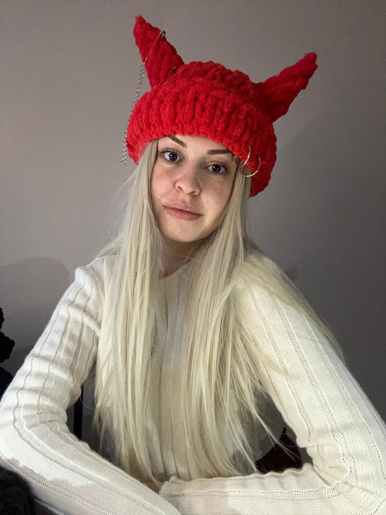 Pode incluir: Um gorro vermelho de croch&ecirc; com chifres de diabo e uma corrente prateada. O gorro &eacute; usado por uma pessoa com cabelo longo e loiro.
