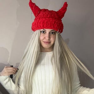 Pode incluir: Um gorro vermelho de croch&ecirc; com chifres de diabo e uma corrente prateada. O gorro &eacute; usado por uma pessoa com cabelo longo e loiro.