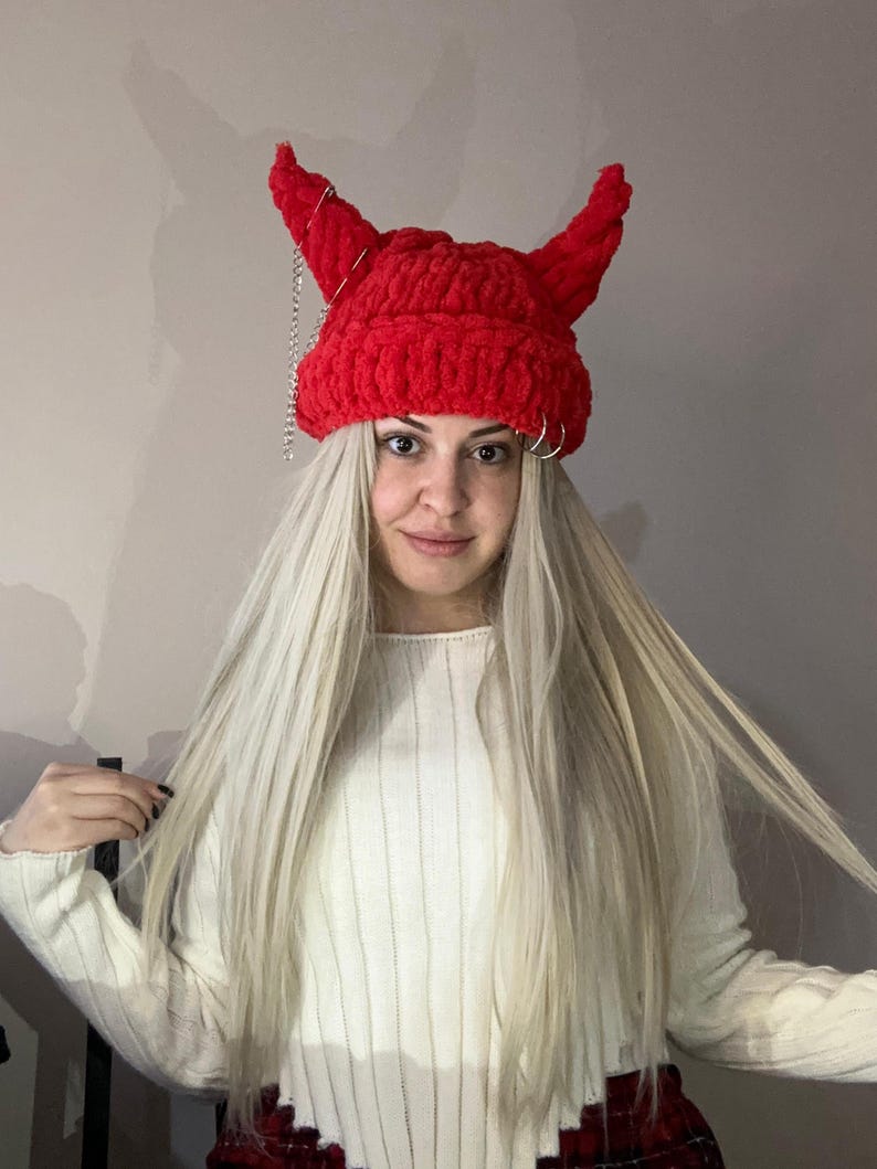 Pode incluir: Um gorro vermelho de croch&ecirc; com chifres de diabo e uma corrente prateada. O gorro &eacute; usado por uma pessoa com cabelo longo e loiro.
