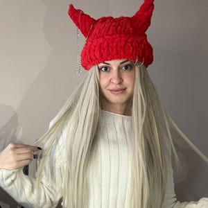 Pode incluir: Boina vermelha de croch&ecirc; com chifres de diabo e uma corrente prateada. A boina &eacute; usada por uma pessoa com cabelo comprido e loiro e um su&eacute;ter branco.