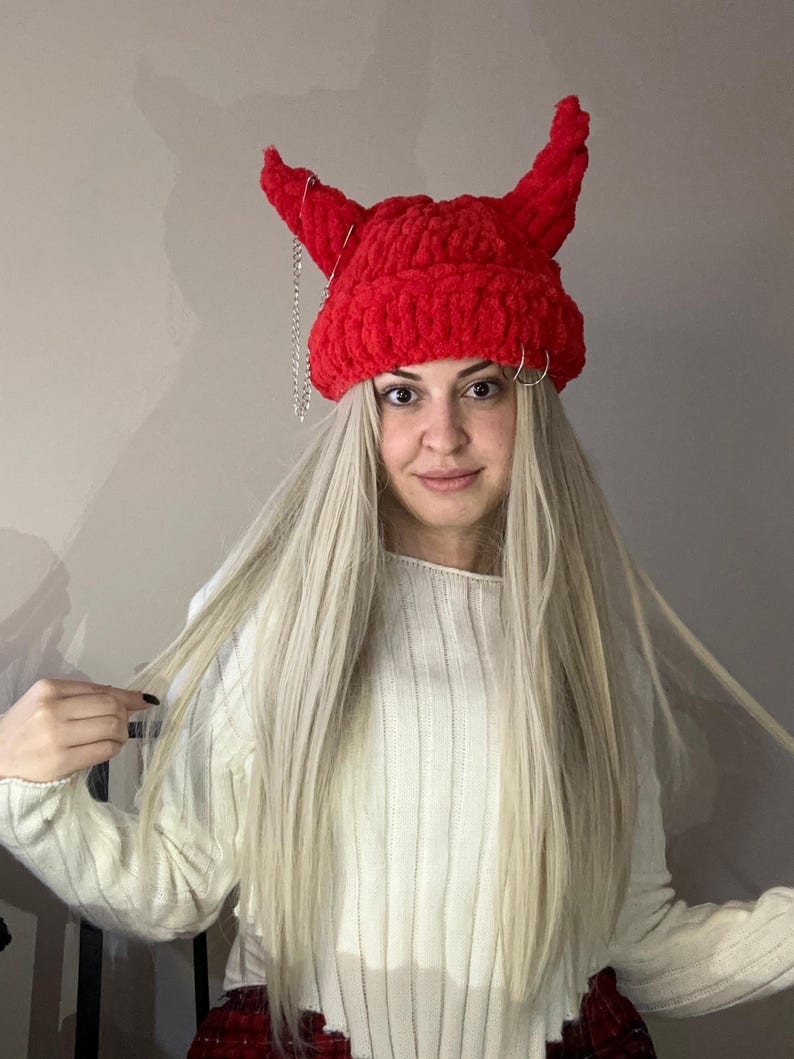 Pode incluir: Boina vermelha de croch&ecirc; com chifres de diabo e uma corrente prateada. A boina &eacute; usada por uma pessoa com cabelo comprido e loiro e um su&eacute;ter branco.