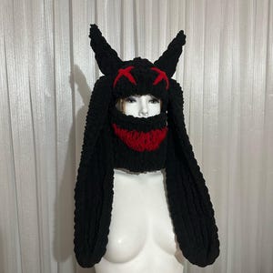 May include: Black knitted hat with long, floppy ears and a red face mask. The hat has a stitched-on design of two red X's on the top.