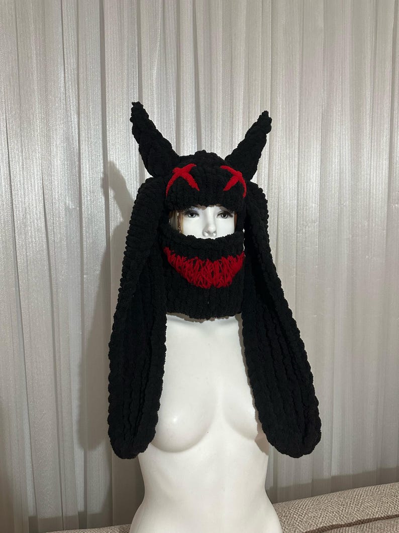 May include: Black knitted hat with long, floppy ears and a red face mask. The hat has a stitched-on design of two red X's on the top.