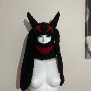 May include: Black knit bunny hat with long ears and a red stitched smile. The hat has red X's on the top.