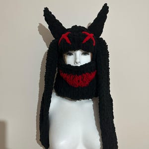 May include: Black knitted hat with long bunny ears and a red knit face mask. The mask has a stitched X design for eyes and a red smile.