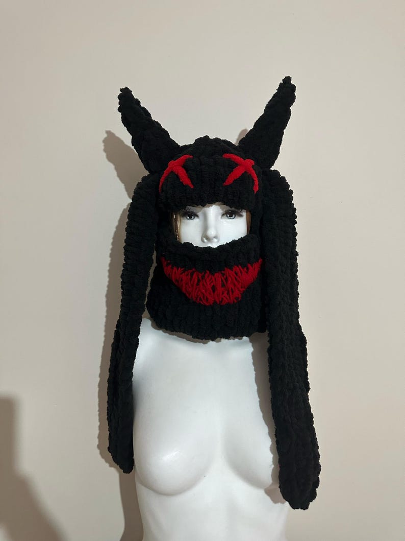 May include: Black knitted hat with long bunny ears and a red knit face mask. The mask has a stitched X design for eyes and a red smile.