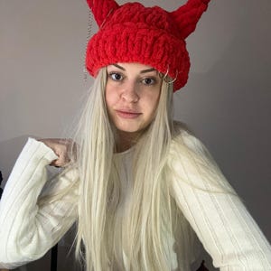 Pode incluir: Um gorro vermelho de croch&ecirc; com chifres de diabo e correntes prateadas. O gorro &eacute; usado por uma pessoa com cabelo comprido e loiro e um su&eacute;ter branco.