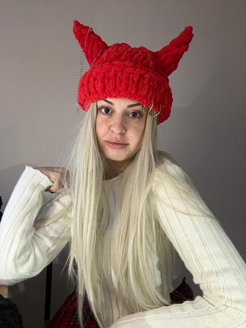 Pode incluir: Um gorro vermelho de croch&ecirc; com chifres de diabo e correntes prateadas. O gorro &eacute; usado por uma pessoa com cabelo comprido e loiro e um su&eacute;ter branco.