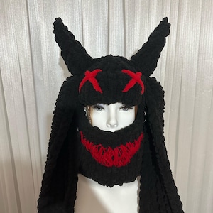 May include: Black knitted hat with long, dangling ear flaps and a red stitched X pattern on the front. The hat has a red stitched smile on the lower face portion.