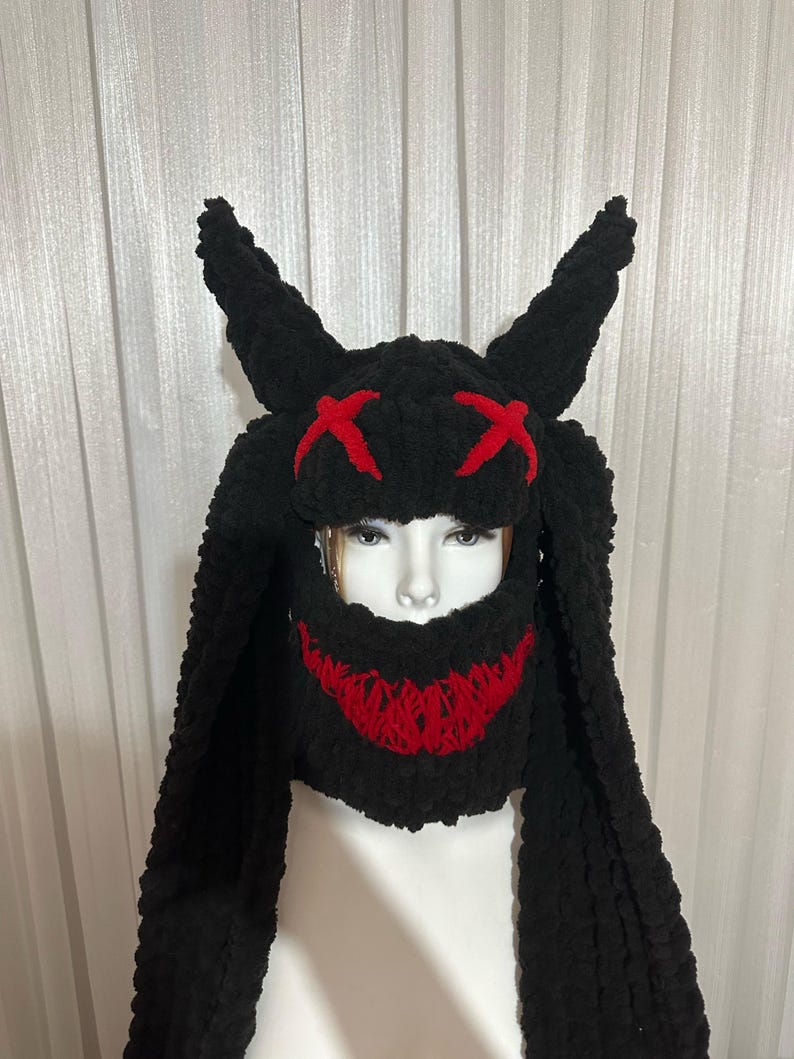 May include: Black knitted hat with long, dangling ear flaps and a red stitched X pattern on the front. The hat has a red stitched smile on the lower face portion.