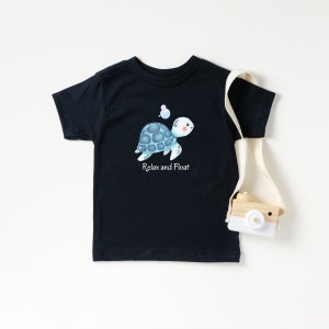 Op de afbeelding: Zwart T-shirt met een cartoon schildpad ontwerp. De schildpad heeft een blauw schild en de woorden "Relax and Float" staan eronder gedrukt. De afbeelding bevat ook een kleine houten speelgoedcamera en een canvas draagtas.
