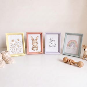 May include: Four rectangular picture frames in pastel yellow, pink, lavender, and light blue. The frames contain prints of wildflowers, a brown bunny, the text "You are loved," and a rainbow. Includes beige knit booties and a wooden rattle.