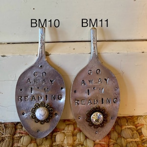 May include: Two silver spoons with the text "Go Away I'm Reading" on them. One spoon has a white stone with a gold trim, the other has a clear rhinestone with a gold trim.