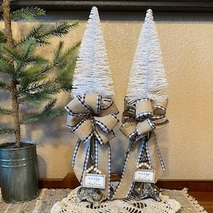 May include: Two white bottle brush Christmas trees with black and white checkered ribbon bows and wooden signs that say "merry christmas". The trees are on a white crocheted doily on a brown woven rug.
