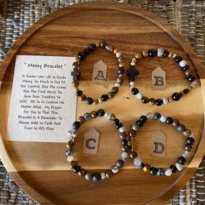 May include: Four beaded bracelets with a cross charm. The bracelets are made with black, brown, and gold beads. The bracelets are on a wooden surface with a note that reads "Messy Bracelet" and a message about faith and trust.