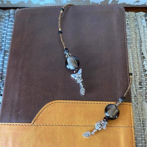 May include: A brown leather-bound journal with a gold and brown beaded bookmark featuring a butterfly and acorn charm.