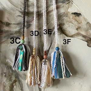 May include: Four handmade tassel pendants with varying designs. One is brown, white, and turquoise with a green bead. The others are white, cream, and blue, each with unique beads and embellishments. The pendants hang against a neutral background.