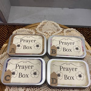 May include: Four rectangular metal boxes with the words "Prayer Box" printed on the labels. Each label has a floral design and a small tag with a letter. The boxes are arranged on a wooden tray with a lace doily.