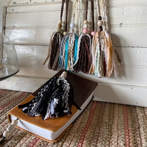 May include: A close-up of a brown leather-bound book with a black and white tassel bookmark. The book is on a woven rug with a brown and white pattern. There are six colorful tassels hanging from the top of the image, each with a wooden bead and a cross charm.