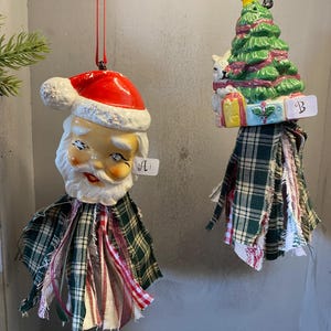 May include: Two Christmas ornaments. One is a Santa Claus head with a red hat and white beard, the other is a green Christmas tree with a yellow star. Both have plaid fabric skirts and are hanging from red and white strings.