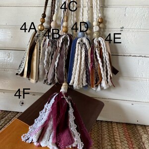 May include: Six different tassel keychains with various colors and textures, including beige, brown, blue, white, and burgundy. Each keychain has a wooden bead and a cross charm. The keychains are labeled 4A, 4B, 4C, 4D, 4E, and 4F.