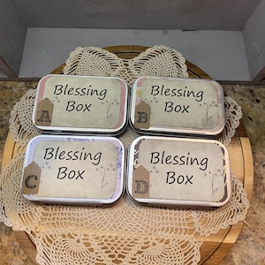May include: Four rectangular metal tins labeled "Blessing Box" with decorative floral accents. Each tin has a different letter tag: A, B, C, and D. The tins are arranged on a wooden tray with a lace doily.