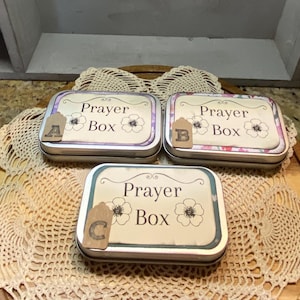 May include: Three rectangular metal prayer boxes with floral designs and the words "Prayer Box" printed on the lids. Each box has a small tag with a letter. The boxes are on a wooden surface with a lace doily.