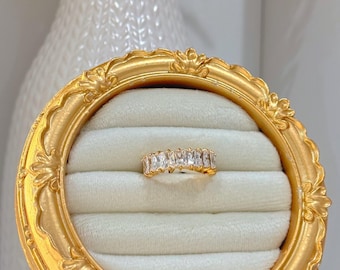 May include: A gold ring with a row of clear baguette-shaped stones set in a gold band. The ring is displayed in a gold ring box with a white velvet interior.