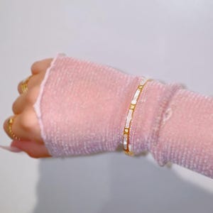 May include: A gold bracelet with rectangular white inlays is worn on a pink sleeve. Several gold rings adorn the fingers. The jewelry is set against a soft, neutral background, highlighting the bracelet's design and the rings.