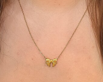 Puede incluir: Un collar dorado con un delicado colgante de lazo. El lazo está intrincadamente diseñado y cuelga de una fina cadena. El collar se muestra sobre un fondo de tono de piel, resaltando los detalles de la joya.