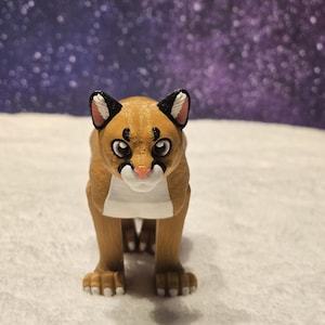 May include: A brown and white 3D printed figurine of a cougar with black markings around the eyes. The figurine is standing on a white surface.