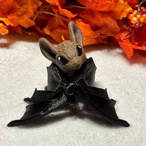 May include: A small brown bat figurine with black wings. The bat is sitting on a white surface with a background of orange fall leaves.
