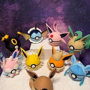 May include: A collection of colorful Pokémon character figurines, including Umbreon, Vaporeon, Sylveon, Jolteon, Flareon, and others. Each figure features distinct colors and designs, with large ears and round bodies. The figurines are arranged on a white surface against a starry background.