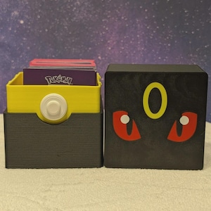 May include: Two 3D-printed storage boxes. One is black with a yellow 'O' and red eyes, the other is yellow and gray with a white button. The yellow box holds cards with a 'Pokemon' logo.