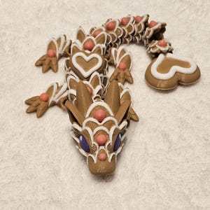 May include: A gingerbread dragon figurine with white icing and pink accents. The dragon has a heart-shaped tail and purple eyes.