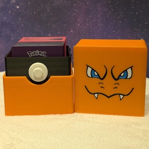 May include: Orange cube-shaped card holder with a cartoon face and blue eyes. The holder is next to a black and orange card holder with a white button. The card holder contains cards with the word "Pokemon" on them.