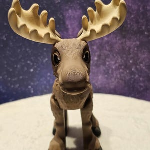May include: A brown moose figurine with large, tan antlers. The moose is standing on a white surface and has a friendly expression.
