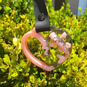 Puede incluir: Colgante en forma de corazón con diseño de pez y rayo. El colgante es de resina rosa y transparente, con una correa y botón negros. Sobre un fondo de arbusto verde.