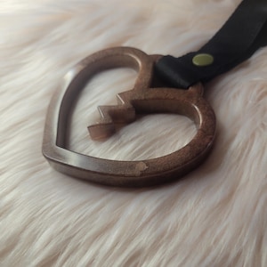 May include: A brown, heart-shaped keychain with a broken design on the bottom left. The keychain has a black strap attached with a brass-colored rivet. The background is a soft, white, fluffy material.