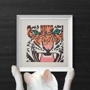 Pode incluir: Um gato branco olha para cima para uma impressão emoldurada de um rosto de tigre. O rosto do tigre é laranja, preto e branco com olhos verdes. A impressão está em uma moldura branca.