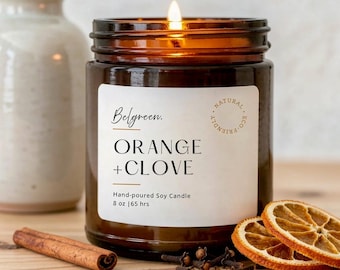 Puede incluir: Una vela de soja encendida en un frasco de vidrio marrón con una etiqueta blanca que dice Belgreen. ORANGE + CLOVE Hand-poured Soy Candle 8 oz | 65 hrs. La etiqueta incluye un sello circular Natural Eco-Friendly. Canela y rodajas de naranja seca.