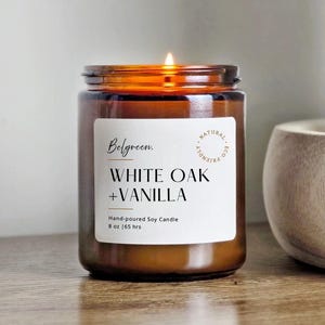 May include: A lit, hand-poured soy candle in a brown glass jar. The label reads "Belgreen. WHITE OAK + VANILLA" with "NATURAL ECO-FRIENDLY" text. The candle is 8 oz and has a 65-hour burn time.