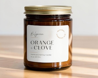 Könnte beinhalten: Eine braune Glas-Kerze mit einem goldenen Metalldeckel und einem weißen Etikett. Das Etikett trägt die Aufschrift Belgreen. ORANGE + CLOVE Hand-poured Soy Candle 8 oz | 65 hrs und einen runden Stempel NATURAL ECO-FRIENDLY.