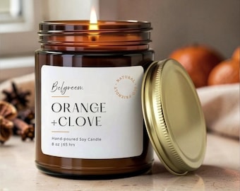 Può includere: Una candela di soia versata a mano in un barattolo di vetro marrone con coperchio dorato. L'etichetta recita "Belgreen. ORANGE + CLOVE" e "Hand-poured Soy Candle 8 oz | 65 hrs". La candela è accesa e c'è un coperchio aperto.